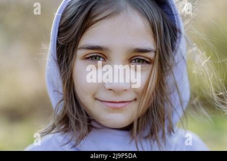 A portrait of pretty child girlin hoodie standing in summer park looking at camera smiling happily. Stock Photo