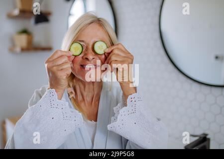 Beautiful senior woman in bathrobe applying cucumber face mask in bathroom, skin care concept. Stock Photo