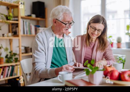 Adult daughter visiting her senior father at home and having coffee together, reading book. Stock Photo