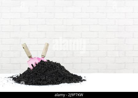 A pile of soil with a pink trowel and fork pushed into it. Isolated on a white surface with a modern white brick background for copy space. Stock Photo