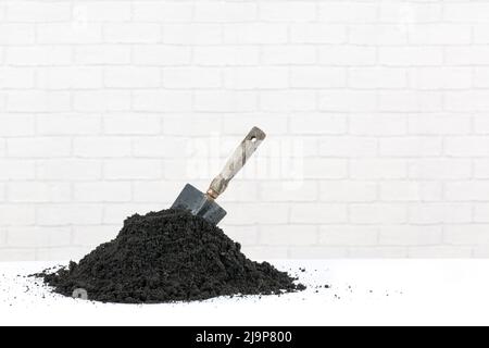 Pile of soil with a wooden handle trowel pushed into it. The earth is on a white surface with a clean white modern brick background. Stock Photo
