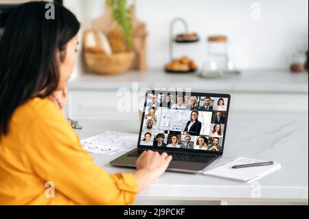 Online learning, professional development. Asian girl, sits at home, listening to a webinar using a video conference, on the screen a group of multiracial people gathered for a brainstorm or lecture Stock Photo