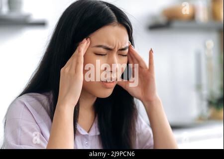 Overworked sad chinese brunette girl, tired of working at a laptop, or experiencing a cold, sitting in the kitchen, suffering of headache, massaging her temples, closing eyes, needs rest and treatment Stock Photo