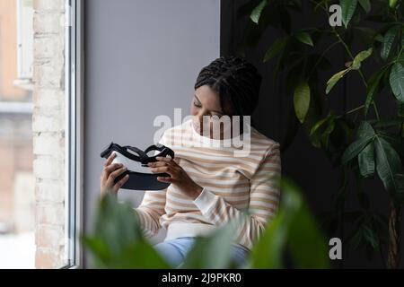 Virtual reality technology. Curious black woman holding VR headset while sitting by window Stock Photo