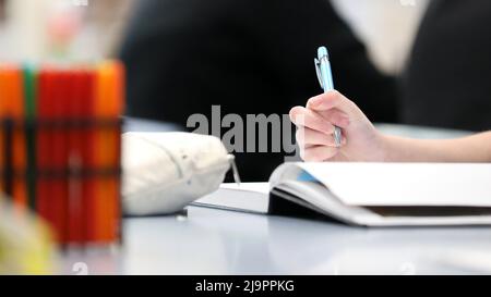 A single hand & fingers holding or gripping a pen about to write or take notes in a blank white open book. Colored markers and pencils case visible. Stock Photo