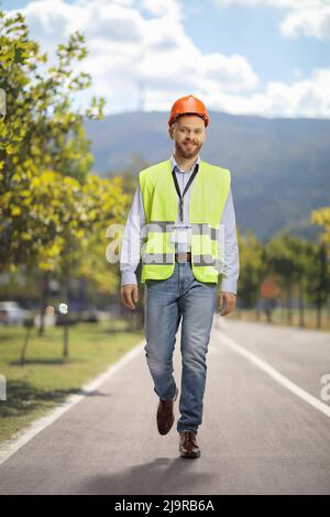 Full length portrait of a site engineer wearing a reflective vest and helmet and walking on a pedestrian lane Stock Photo