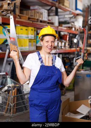 Young female in uniform and helmet holding saw near shelves with tools Stock Photo