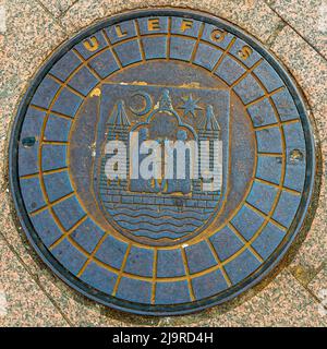 A manhole cover set into the pavement in Aarhus, Denmark, shows the city's coat of arms: two figures, a crescent moon and a star above an anchor. The rim reads "Ulefos", after the Norwegian ironworks Ulefos Jernverk, founded in 1657 in the town of Ulefoss, which still supplies manhole covers to cities across Scandinavia today. Stock Photo