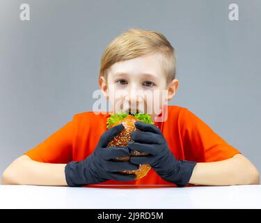a boy sitting at a table holds a juicy fried burger in front of him with a salad inside, he wants to take a bite of it Stock Photo