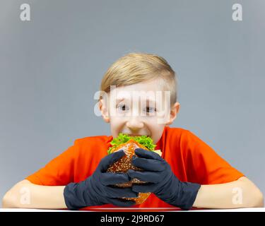 A cheerful boy sits at a table and holds a juicy burger in front of him Stock Photo
