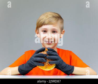 cheerful smiling boy sits at the table and holds a juicy burger in front of him Stock Photo