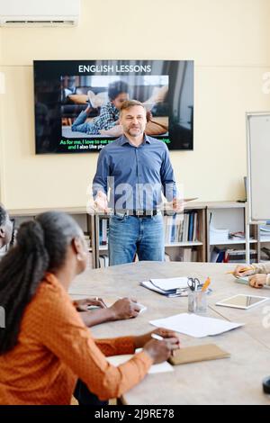 Vertical shot of mature Caucasian man standing at desk in front of multi-ethnic students teaching them English language Stock Photo