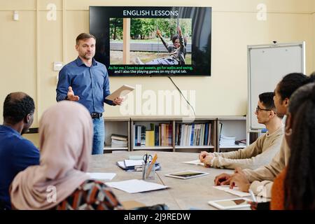 Professional English language teacher standing at table in front of multi-ethnic migrant students explaining something to them Stock Photo