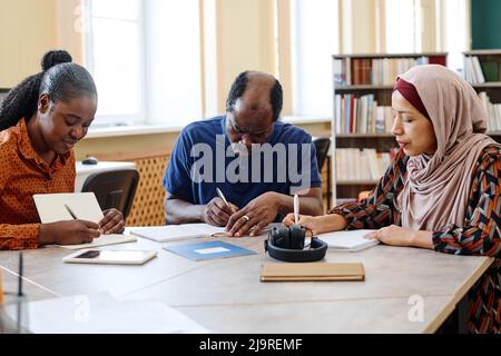 Group of three multi-ethnic immigrant students sitting at table in library doing writing task during lesson Stock Photo