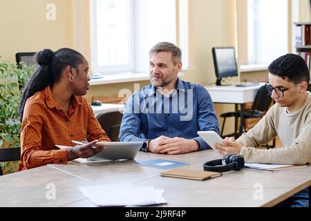 Mature Caucasian man sitting at table with his immigrant students talking to them during English language class Stock Photo