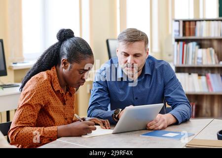 Mature Caucasian teacher sitting next to his Black student explaining her how to do task and showing something on digital tablet Stock Photo