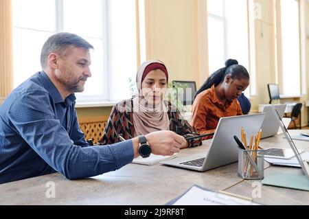 Mature Caucasian teacher holding pen showing something on laptop screen and explaining it to Muslim woman in hijab during lesson Stock Photo