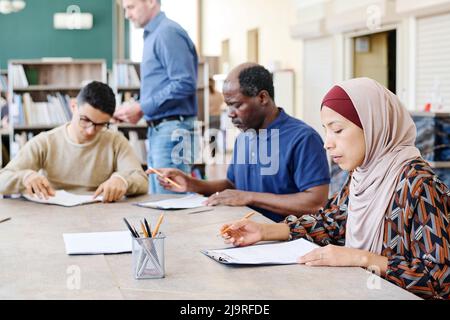 Group of multi-ethnic immigrant students sitting at table in library working on English language test Stock Photo