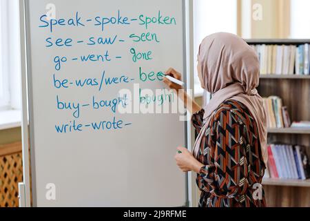 Unrecognizable woman wearing hijab standing at whiteboard doing task with irregular verbs during English language for immigrants lesson Stock Photo