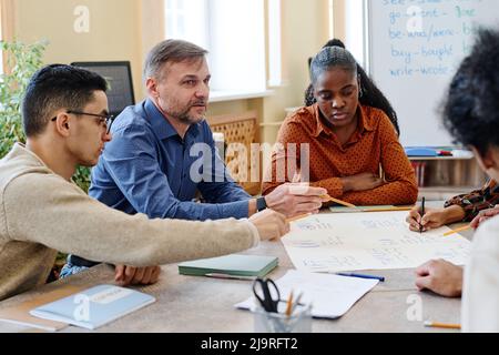 Mature teacher and his immigrant students working on educational English language poster together in group Stock Photo