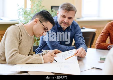 Mature Caucasian teacher of English language watching Middle Eastern immigrant student writing something on poster Stock Photo