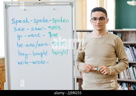 Horizontal medium portrait of young Middle-Eastern student standing at whiteboard with irregular verbs written on it looking at camera Stock Photo