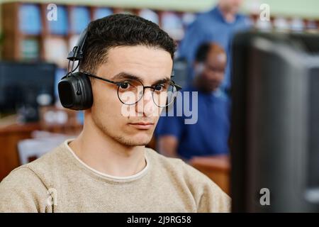 Young serious Middle Eastern studio wearing eyeglasses and headphones doing listening task during English lesson Stock Photo