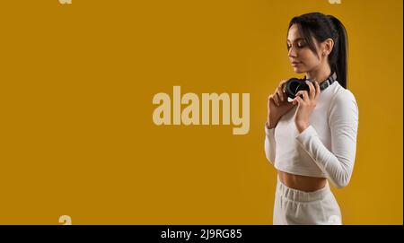 Side view of brunette girl with ponytail standing, looking down. Pretty slim young female wearing white top and trousers, holding earphones on neck. Concept of modern life. Stock Photo