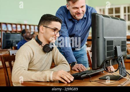 Cheerful mature teacher watching task his student finishing doing using desktop computer in library Stock Photo