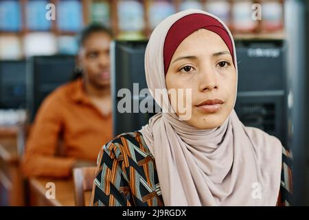 Portrait of young Muslim woman wearing hijab sitting in university library looking at computer screen Stock Photo