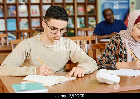 Serious young Middle Eastern man sitting at desk in classroom working on essay in notebook Stock Photo