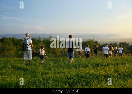 Back view of group of kids with old man going forward in hills. Children with teacher with gray hair hiking, traveling, walking, holding by hands. Concept of importance of support. Stock Photo