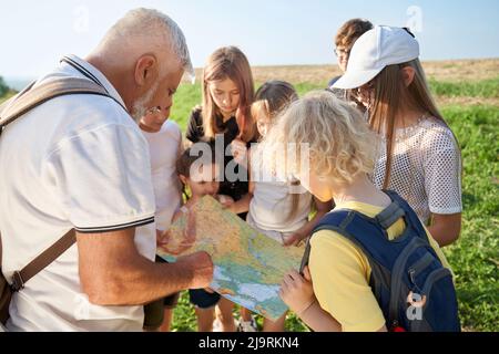 Close up of old man wearing white T-shirt and rucksack, showing map to children outside. Kids looking at map, searching route, camping in hills. Concept of mount hiking. Stock Photo