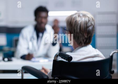 Selective focus on sick senior patient with disability talking with physician about illness examination appointment. Woman in wheelchair conversating with doctor about health check and prescribed drugs Stock Photo