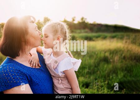 Grandmother holding granddaughter on her arms and kissing nose. Happy childhood, family love concept. Support and togetherness. Two people outdoor with sunlight background. High quality photo Stock Photo