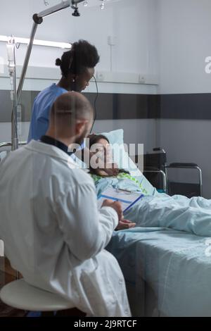 Doctor during morning rounds checking clipboard with patient records while nurse is putting oxygen mask on unconscious woman. Caregiver attending recovering female in modern hospital ward. Stock Photo