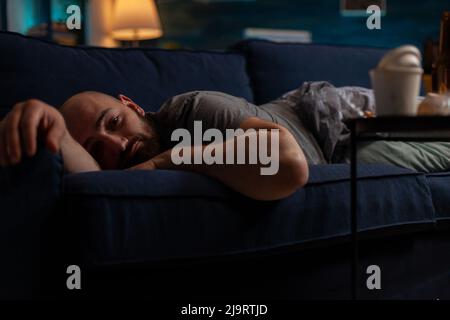 Depressed anxious adult with mental illness laying on couch, feeling pensive and frustrated. Sad desperate man with suicidal thoughts resting on sofa, dealing with chronic disease difficulties. Stock Photo