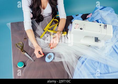 brunette businesswoman seamstress tailor dressmaker works in the atelier with sewing machine and measuring tape on a blue background in the studio. Stock Photo