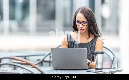 Brunette with glasses works outdoors surrounded by office buildings on a laptop. Stock Photo