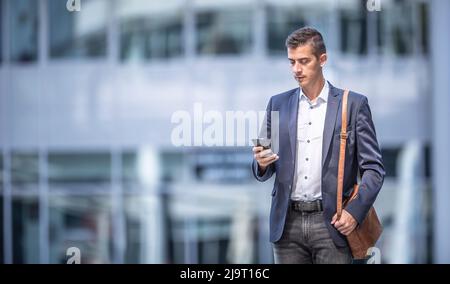 Businessman in a casual outfit wears mens' purse, standing in front of an office building, chekcing his cell phone. Stock Photo