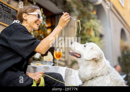 Woman eating pasta with her cute white dog at the restaurant Stock Photo