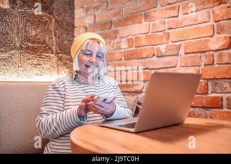 Close-up portrait of smiling senior mature middle aged businesswoman using phone and laptop working and web surfing on desk at cafe Stock Photo