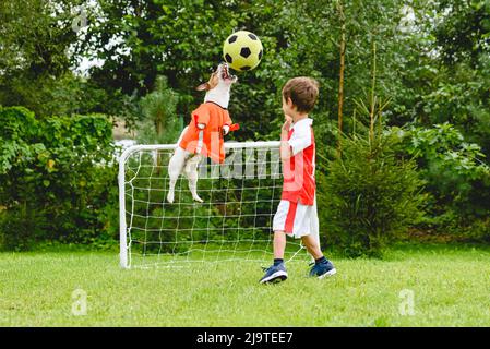 Sportive boy and his dog both wearing soccer kit playing with football ball at park playground outdoors on summer day Stock Photo