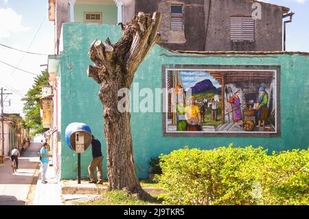 Incidental Cuban people are seen by a small park in the Colon street. A man speaks on an Etecsa payphone. A tile painting or art decorates the lateral Stock Photo