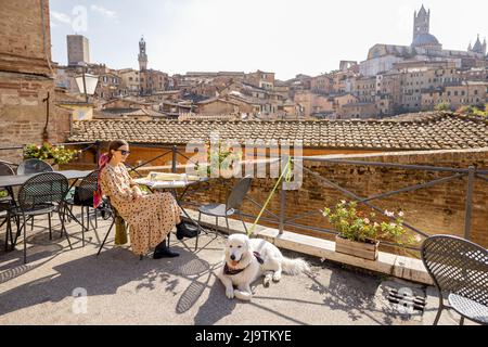 Young woman having lunch, sitting with a dog at outdoor restaurant in Siena town Stock Photo