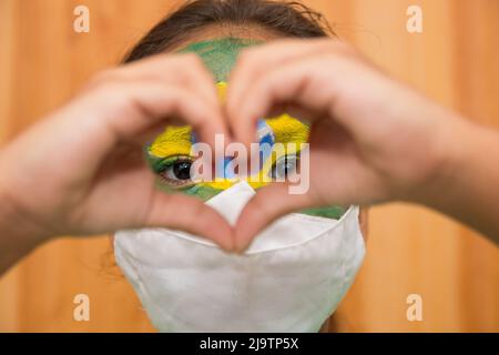 child with mask with his face painted with the flag of brazil, making a heart with his hands. Stock Photo