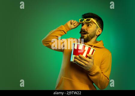Portrait of young man in glasses with popcorn box isolated over green background in neon light. Shocking movie scene Stock Photo