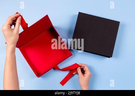 Female hands opening empty red elegant luxury box with ribbon. Mockup present gift boxes on light blue background. Top view Stock Photo