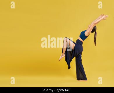 Modern female dancer wearing wide slits pants, performing dance poses and movements outdoors. Yogi girl practicing yoga, with copy space, isolated on orange studio background. Concept of lifestyle. Stock Photo