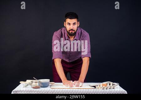 young and attractive Italian man baker with beard makes the dough sprinkled with flour Italian pizza on a black background in the studio Stock Photo